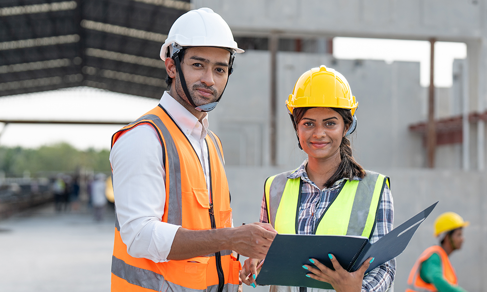 Ein Bauingenieur und eine Bauingenieurin asiatischer Herkunft, die Schutzweste und Helm tragen, auf einer Baustelle.